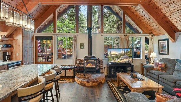 Interior acogedor de una cabaña con cocina, comedor y sala de estar, destacando un gran techo de madera, una chimenea y ventanas amplias.
