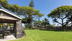 A covered outdoor seating area with stone pillars, a volleyball net, and a lawn with trees and a stone wall. - Surf & Racquet Club #24 New Beds, New You! (Kailua-Kona)