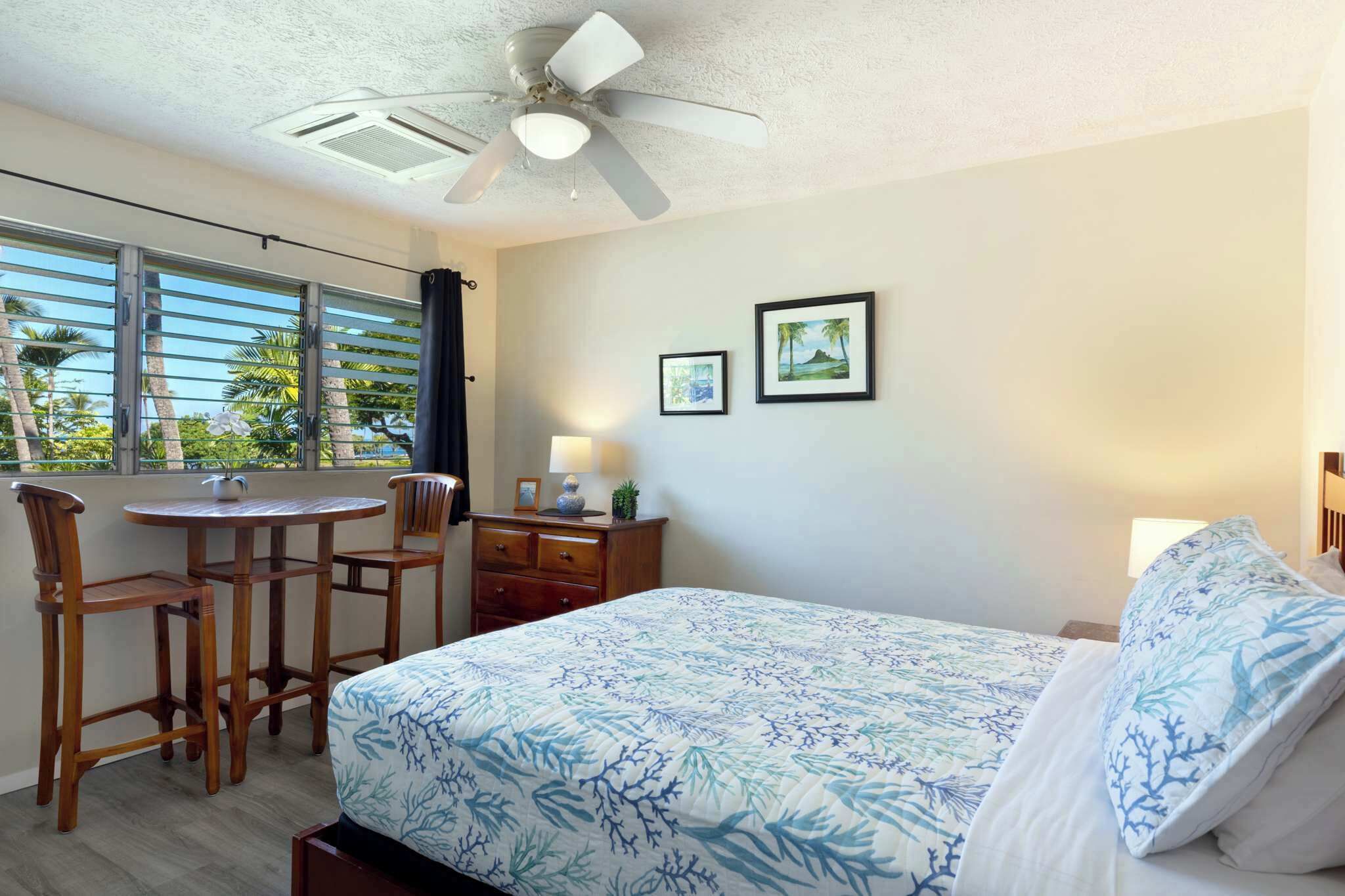A bedroom with a bed, a desk, two chairs, a ceiling fan, and a window with blinds.