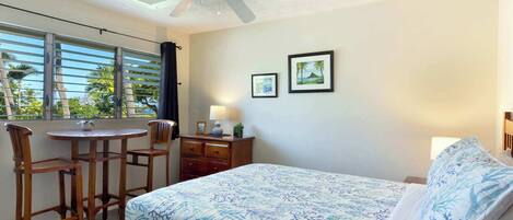 A bedroom with a bed, a desk, two chairs, a ceiling fan, and a window with blinds.
