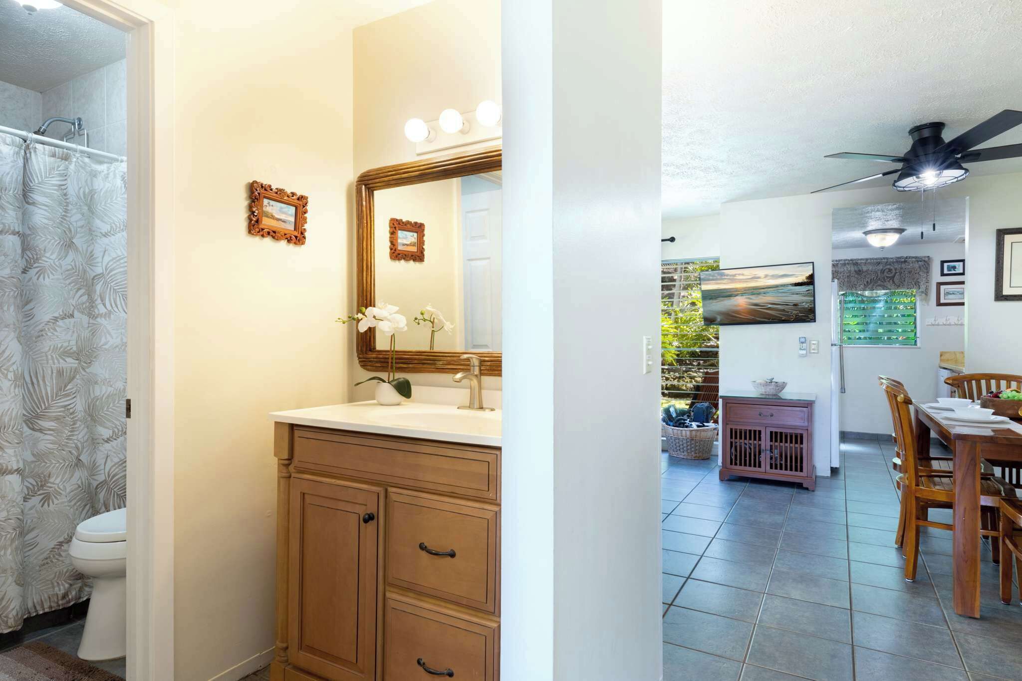 A bathroom with a sink, mirror, and wooden cabinets. There is a shower curtain with a leaf pattern. The bathroom connects to a dining area with a table and chairs.