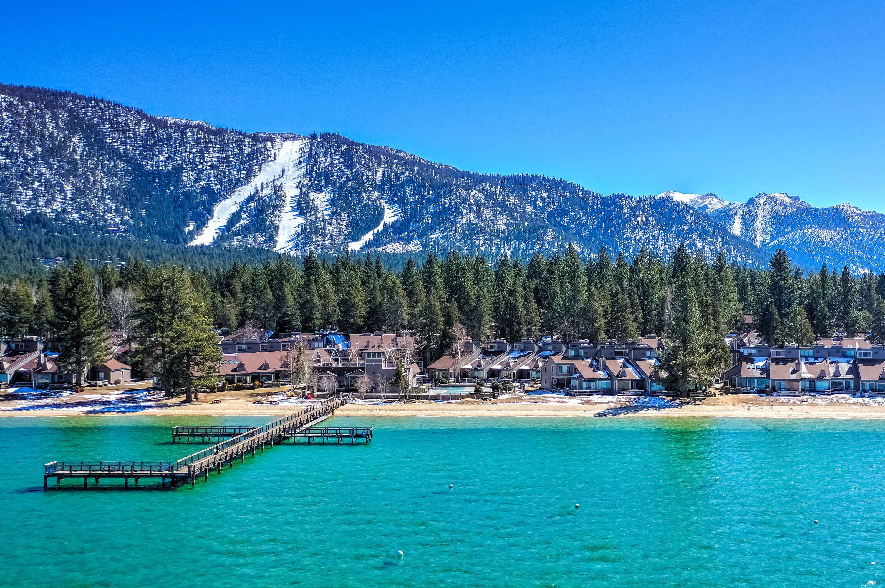 A coastal resort with a wooden pier extending into turquoise waters, surrounded by snow-capped mountains and dense forests.