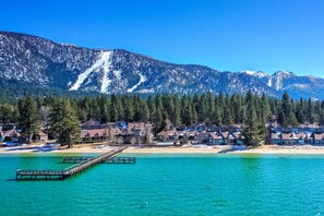 A coastal resort with a wooden pier extending into turquoise waters, surrounded by snow-capped mountains and dense forests.