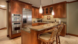A kitchen with wooden cabinets, stainless steel appliances, a granite countertop, and a central island with bar stools. - Kolea 11D Walk to the Beach (Waikoloa)