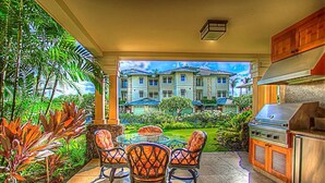 An outdoor kitchen area with a grill, surrounded by tropical plants and a view of a multi-story building. - Kolea 11D Walk to the Beach (Waikoloa)