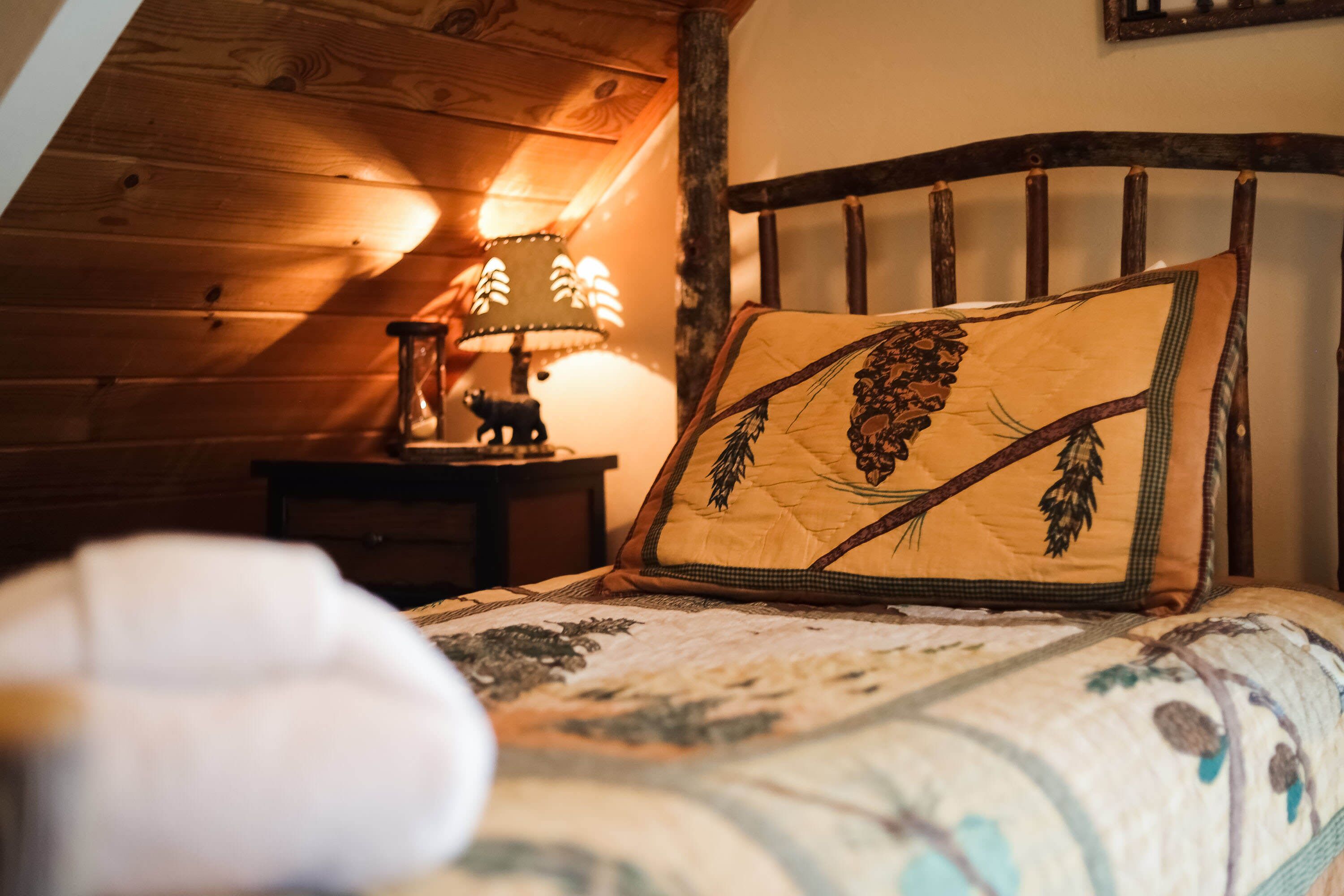 Un dormitorio acogedor con techo de madera, una cama con colcha estampada de flores, una mesita de noche con lámpara y una figura de un oso, y un marco de fotos colgado en la pared.