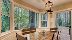 Un espace repas comprenant une grande table et des chaises en bois, de grandes fenêtres donnant sur la forêt et un lustre.