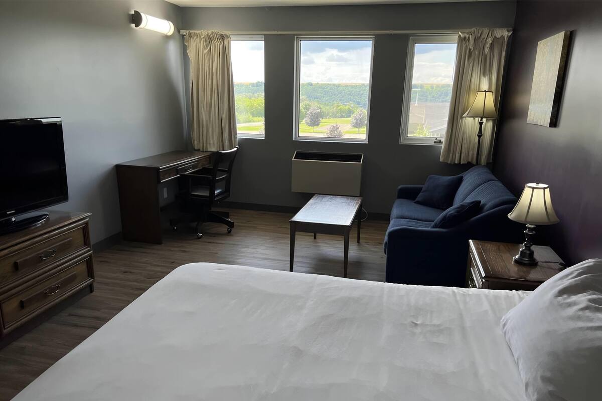 A hotel room with a bed, a desk, a chair, a television, and two windows with curtains.