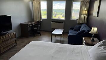 Et hotelværelse med en seng, et skrivebord, en stol, et fjernsyn og to vinduer med gardiner.