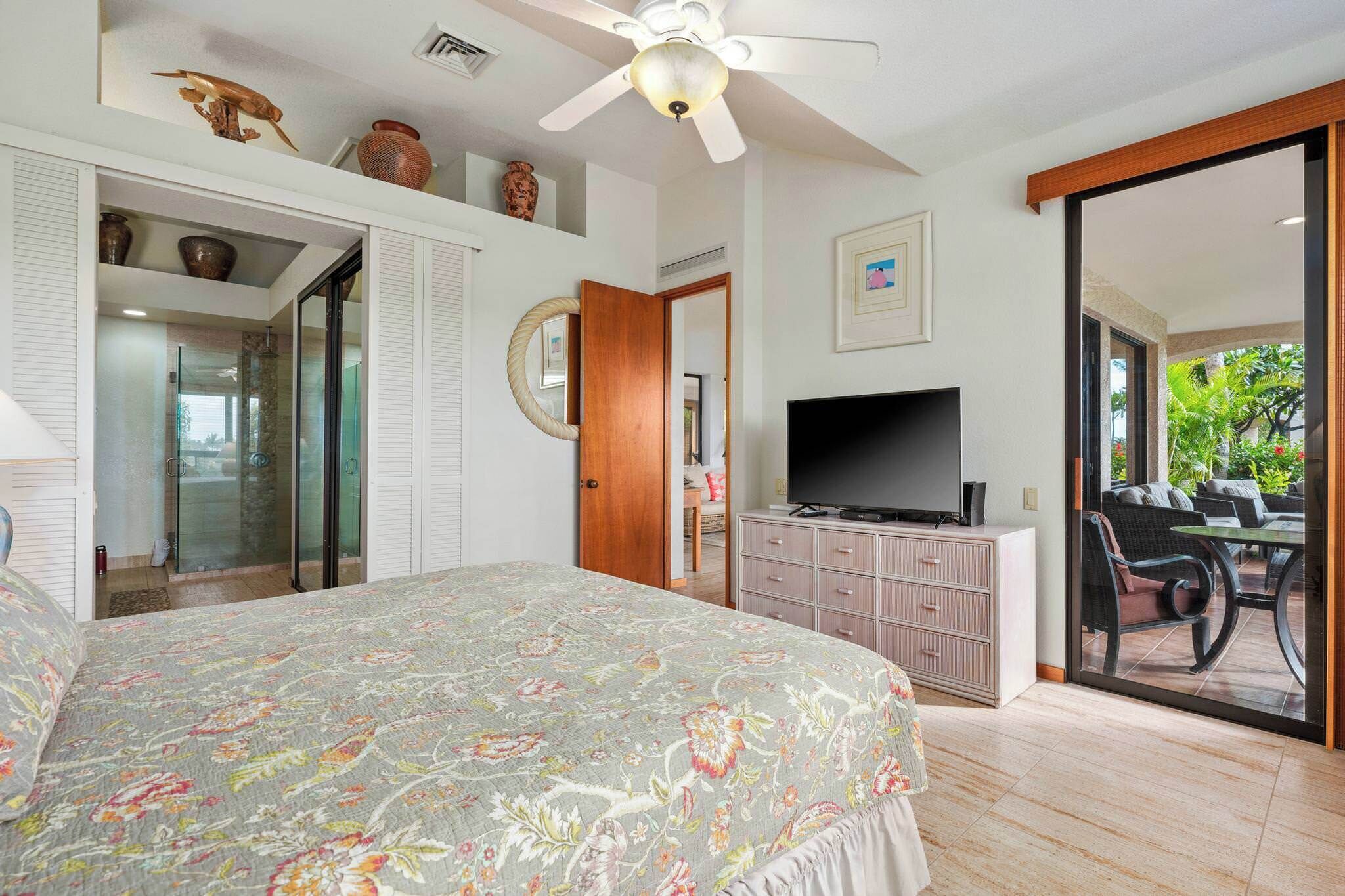 A bedroom with a bed, a TV, a desk with a chair, and a sliding glass door leading to a balcony.