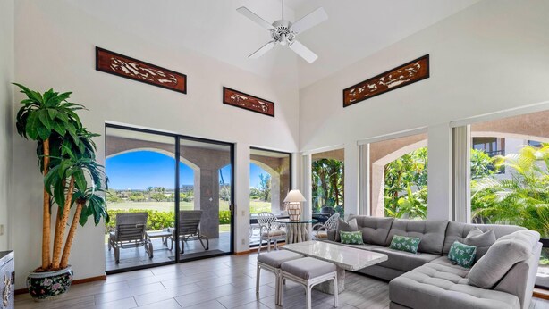 A spacious living room with a grey sectional sofa, a white coffee table, and a potted plant. There are sliding glass doors leading to a balcony with outdoor furniture. - Exclusive Single-story Shores Golf Villa 20 - 3 Ensuite (Waikoloa)