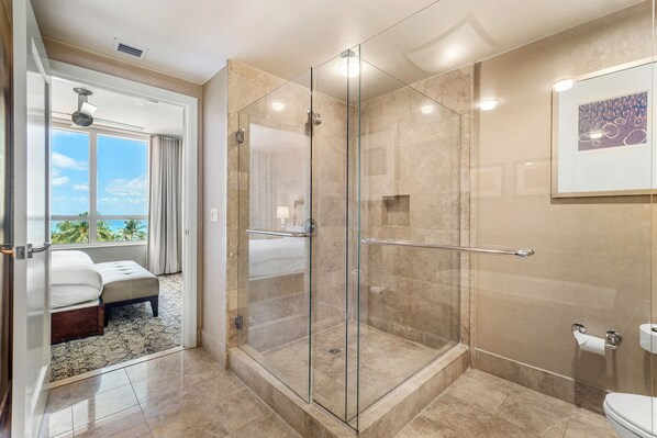 A modern bathroom with a glass-enclosed shower, a toilet, and a large window offering a view of the sky and palm trees.