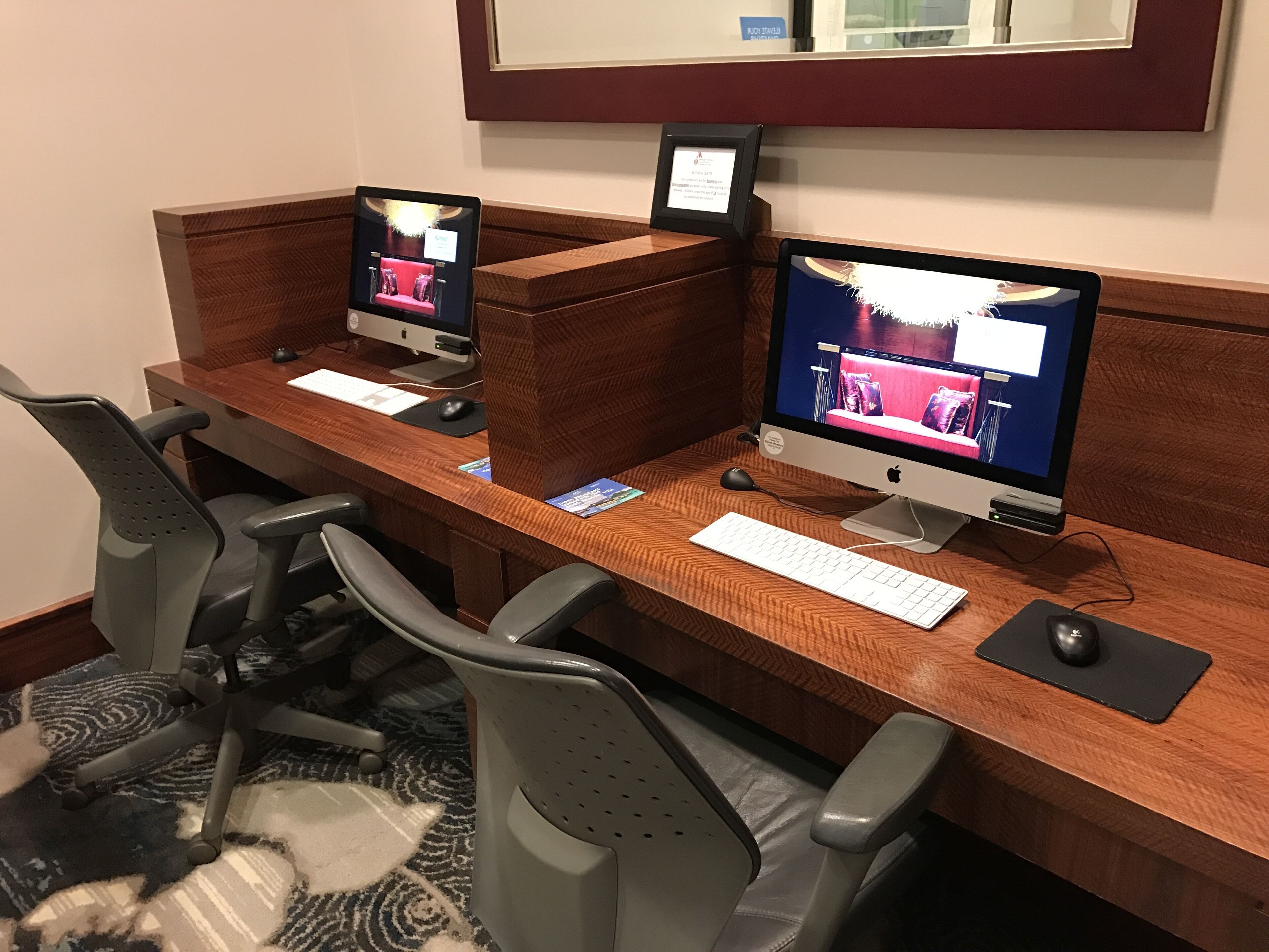 A hotel workspace with two Apple iMac computers, a white keyboard, and grey office chairs.