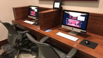 A hotel workspace with two Apple iMac computers, a white keyboard, and grey office chairs.