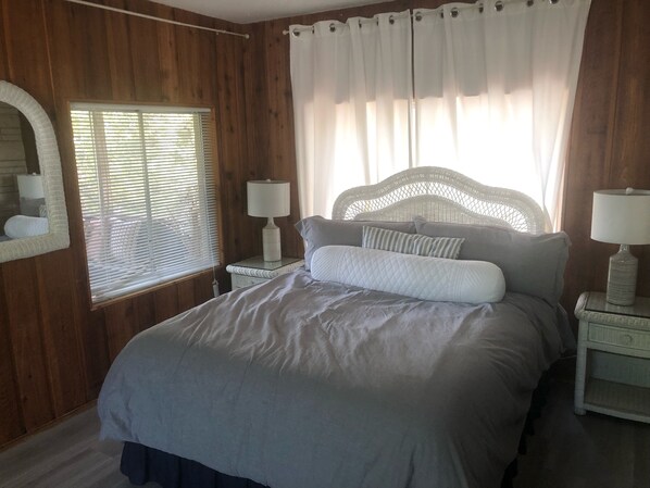 A bedroom with a wooden wall, a bed with a white quilt, two bedside tables with lamps, and a window with blinds.