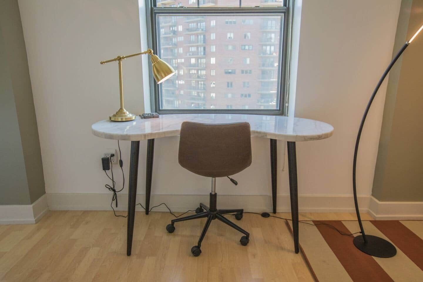 A corner office with a white desk, a brown office chair, a gold desk lamp, and a view of a building through a window.