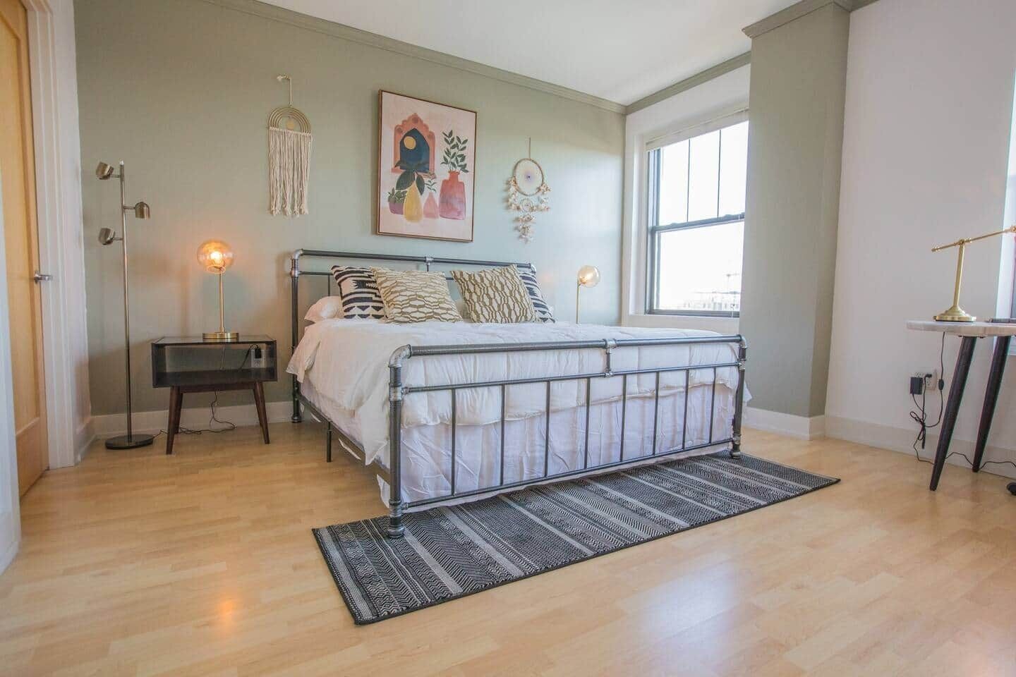 A bedroom with a metal bed frame, a bedside table, a floor lamp, a hanging macrame decoration, and a wall art piece.