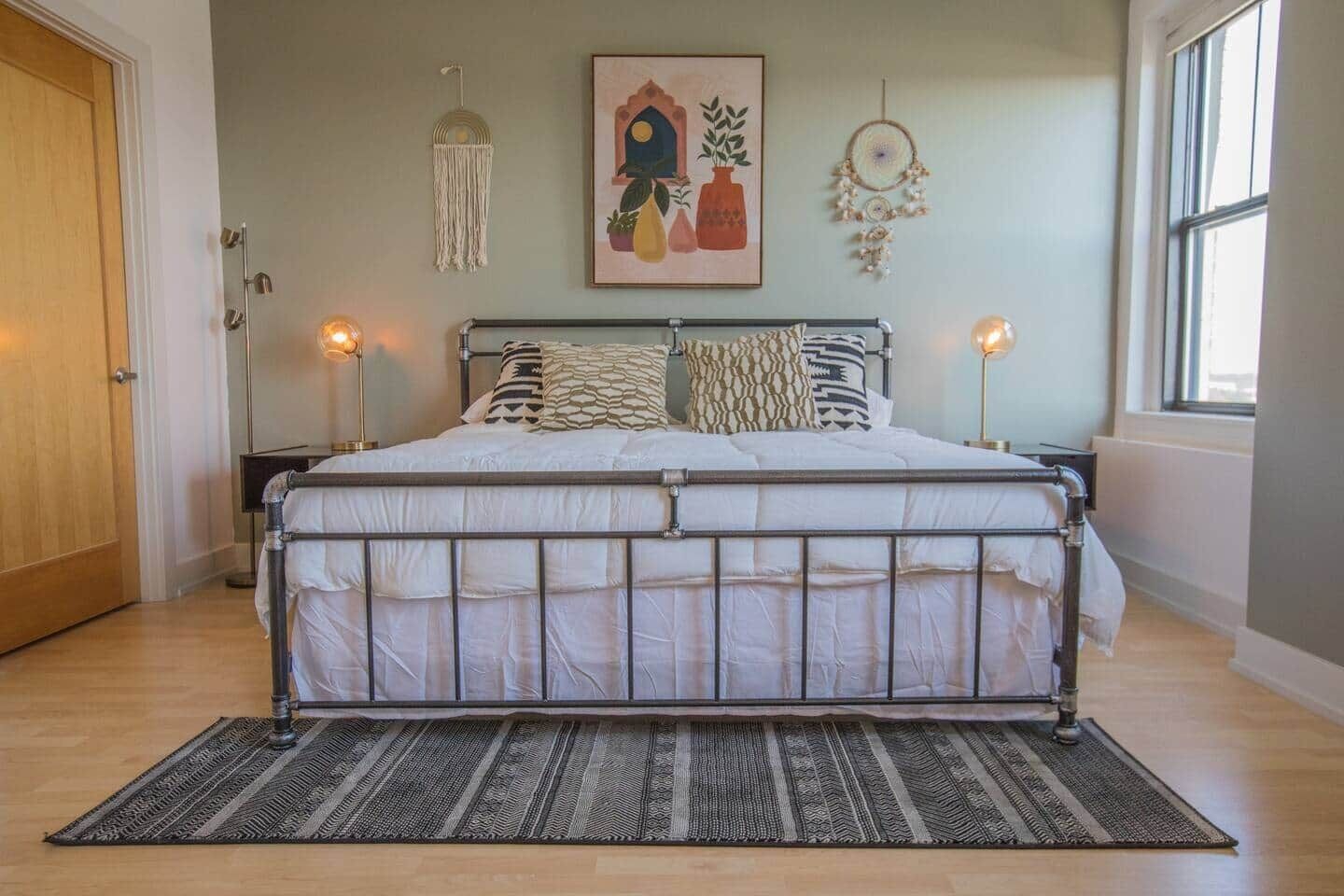 Una cama doble con estructura de metal, un tapete estampado y una pared decorada con un atrapasueños y un cuadro.