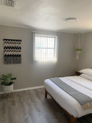 Ein Schlafzimmer mit einem Bett, einem Fenster mit Jalousien, einem Wandbehang aus Makramee, einer hängenden Makramee-Pflanze und einer Topfpflanze.