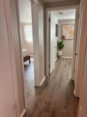 A hallway with wooden flooring, white walls, and a door leading to a bedroom with a bed and window blinds. - Boho Chic Home (New port richey)