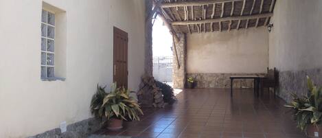 A covered patio area with a rustic wooden ceiling, a stone wall, and a tiled floor. There is a wooden door and a small window on the left.