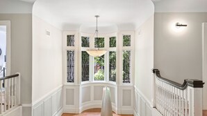 A hallway with a white railing, a hanging light fixture, and a view through a bay window with decorative glass panes.