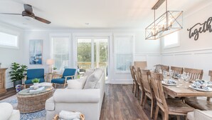 Un salon spacieux avec un canapé blanc, des fauteuils bleus, une table à manger dressée pour six personnes et une grande fenêtre équipée de stores.