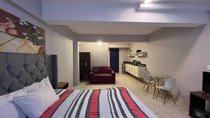 Een slaapkamer met een groot bed, een rode bank, een kleine eettafel met stoelen, een kitchenette en een deur naar een andere kamer.