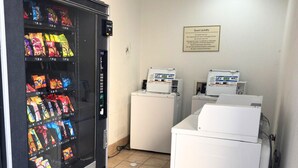 Distributore automatico con snack, cartello per la lavanderia degli ospiti e un piccolo elettrodomestico bianco, probabilmente una macchina per il caffè, in una stanza con pavimento piastrellato.