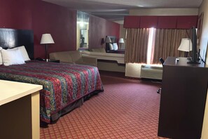Ein Hotelzimmer mit einem großen Bett, einem Schreibtisch, einem Sessel, einem Spiegel und einem Fenster mit Vorhängen.
