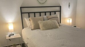 Una cama bien hecha con edredón blanco y almohadas, una mesita de noche con lámpara y un adorno en la pared.