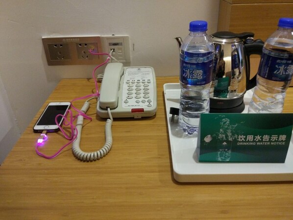 Escritorio de habitación de hotel con teléfono fijo, un teléfono móvil cargando, dos botellas de agua, una tetera y un cartel informativo sobre el agua.