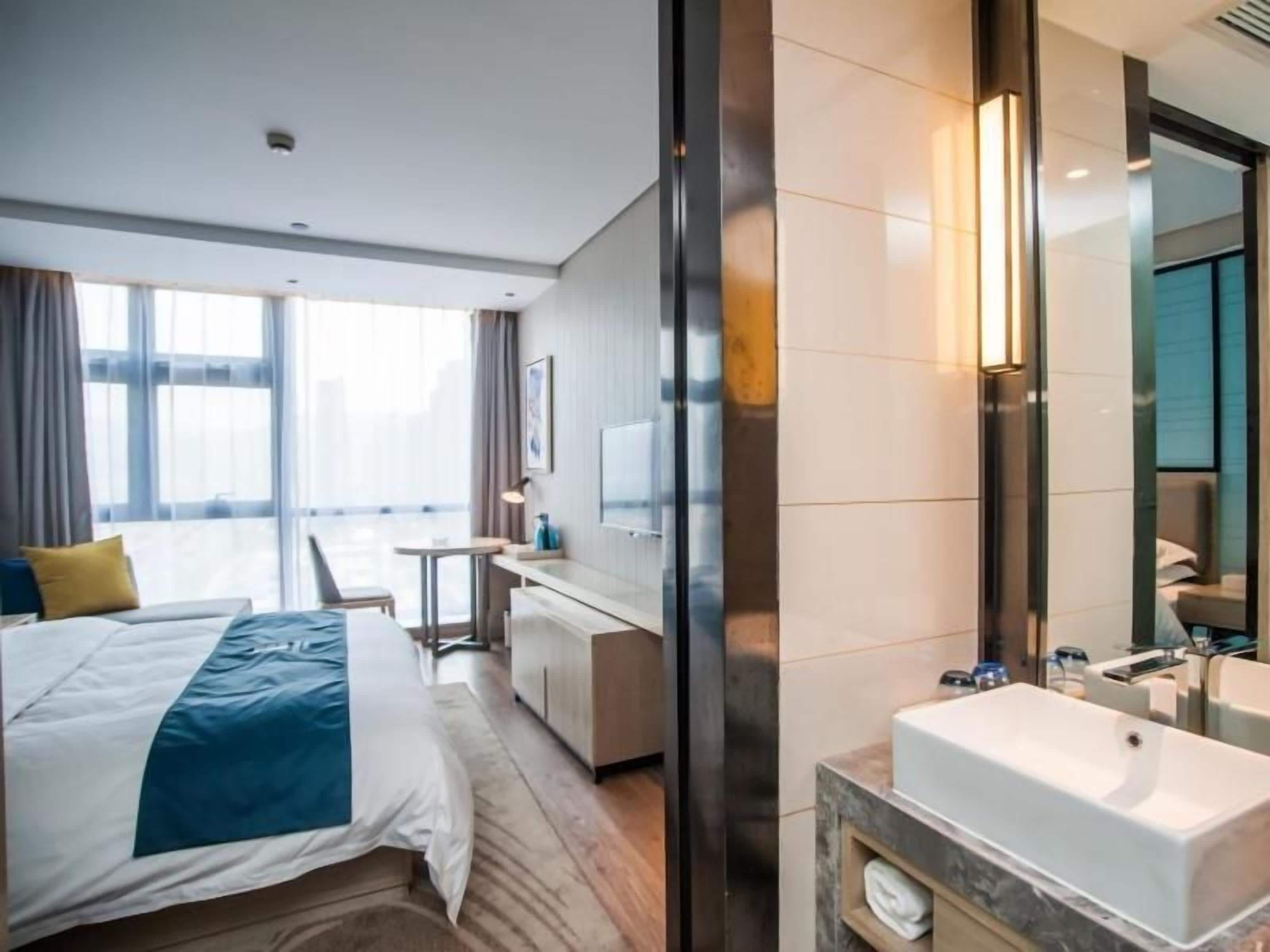 Habitación de hotel con cama, escritorio, baño con lavamanos y espejo, y ventanas grandes.