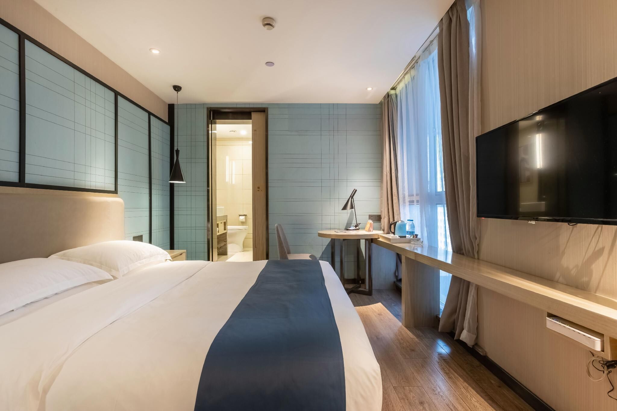 A hotel room with a large bed, a desk with a lamp, a TV, and a bathroom visible through an open door.