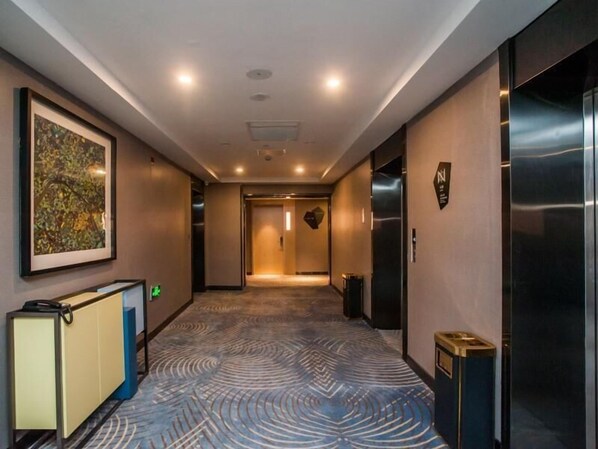 A modern hotel corridor with a patterned carpet, a reception desk, an elevator, and a framed artwork on the wall.