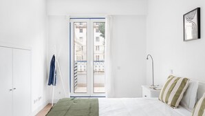 A bedroom with a bed, a nightstand, a lamp, a window with a view of buildings, and a door. - JOIVY Modern flat w/river view near Bica Elevator (Lisboa)