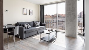 Un salon moderne avec un canapé gris, une table basse en verre et un coin repas comprenant une chaise et une table. De grandes fenêtres offrent une vue sur les bâtiments et le paysage urbain.