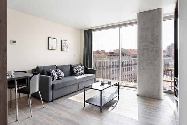 A modern living room with a grey sofa, a glass coffee table, and a dining area with a chair and table. Large windows offer a view of buildings and a cityscape. - JOIVY Amazing central studio w/river view in Lisbon (Lisboa)