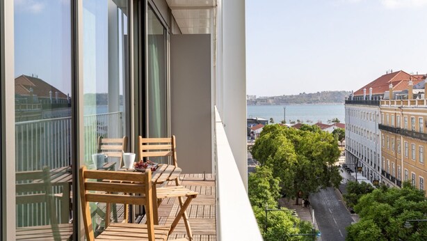 Un balcon avec des chaises et une table en bois, donnant sur un paysage urbain avec des bâtiments et une étendue d’eau.