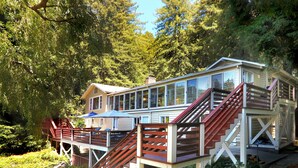 A wooden house with a deck and stairs, surrounded by trees. - River Queen: Historic 5 Bed/5.5 Bath, Dock, Boats + New Hot Tub! (Monte Rio)