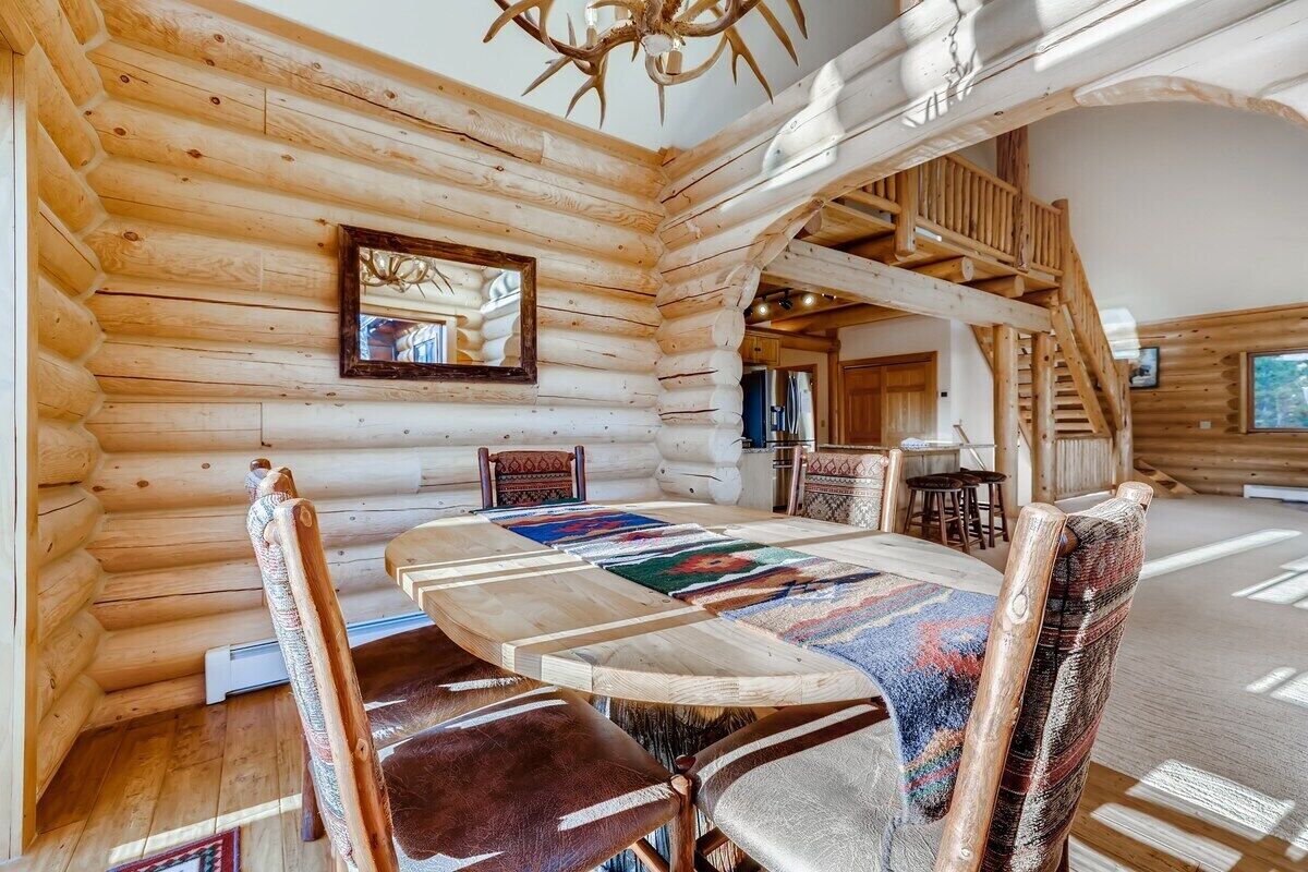 Interior de una cabaña de troncos con una mesa de comedor, sillas, una araña y una escalera.
