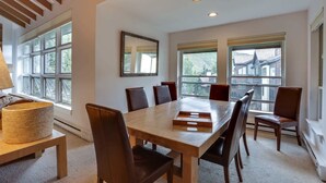 A modern dining room with a wooden table and chairs, large windows, and a view of buildings outside.