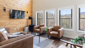 A modern living room with a wooden wall, a flat-screen TV, a fireplace, and large windows offering a view of trees and mountains.
