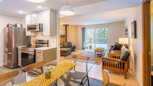Uma cozinha moderna com eletrodomésticos de aço inoxidável, um espaço de refeições com mesa de vidro e uma sala de estar com sofá e mesa de centro.