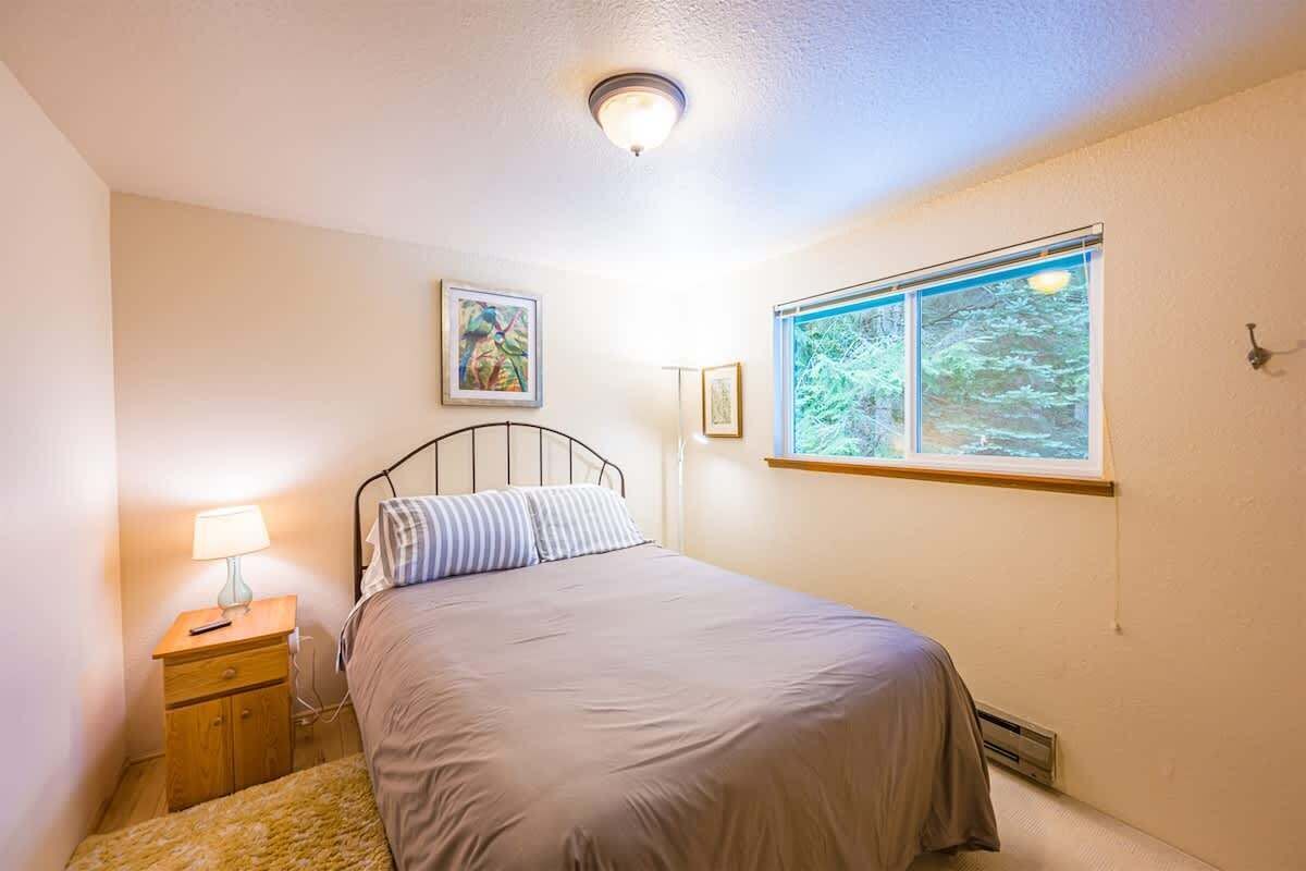 A bedroom with a bed, a nightstand, a lamp, a window with a view of trees, and a ceiling light.