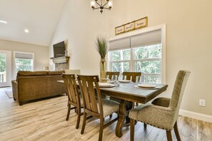 A dining area with a wooden table, chairs, and a vase. There is a sofa and a TV in the background. - Dream Ridge | Ellijay, GA (Ellijay)