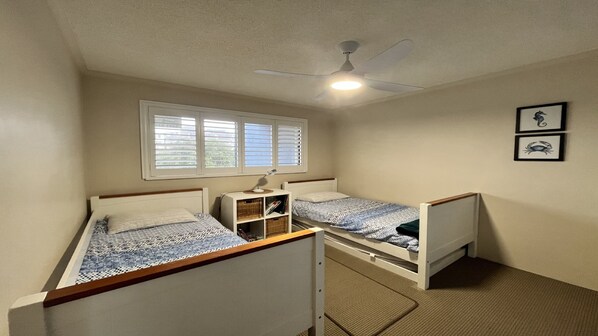 A bedroom with two single beds, a ceiling fan, a window with blinds, and a small desk with a lamp.