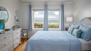 A bedroom with a large bed, a dresser, a chair, and a view of the beach through the window. - 'Pop's Inn" - Oceanfront Home, 4 Bedrooms / 2 Bathrooms (Holden Beach)