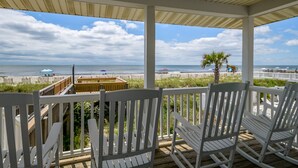 A covered deck with white railings and rocking chairs overlooking a beach with umbrellas and a palm tree. - 'Pop's Inn" - Oceanfront Home, 4 Bedrooms / 2 Bathrooms (Holden Beach)