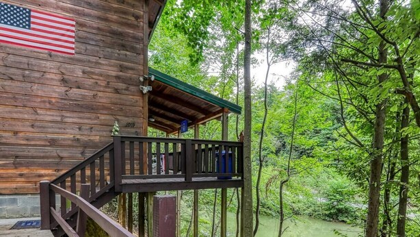 A wooden cabin with a balcony overlooking a river, surrounded by dense forest. - Secluded Smokies Cabin | Private Pond + Hot Tub + King Bed (Sevierville)
